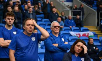 Disheartened Cardiff City fans react in the stands of their home stadium, with somber expressions and heads in hands, following the club’s relegation to League One.